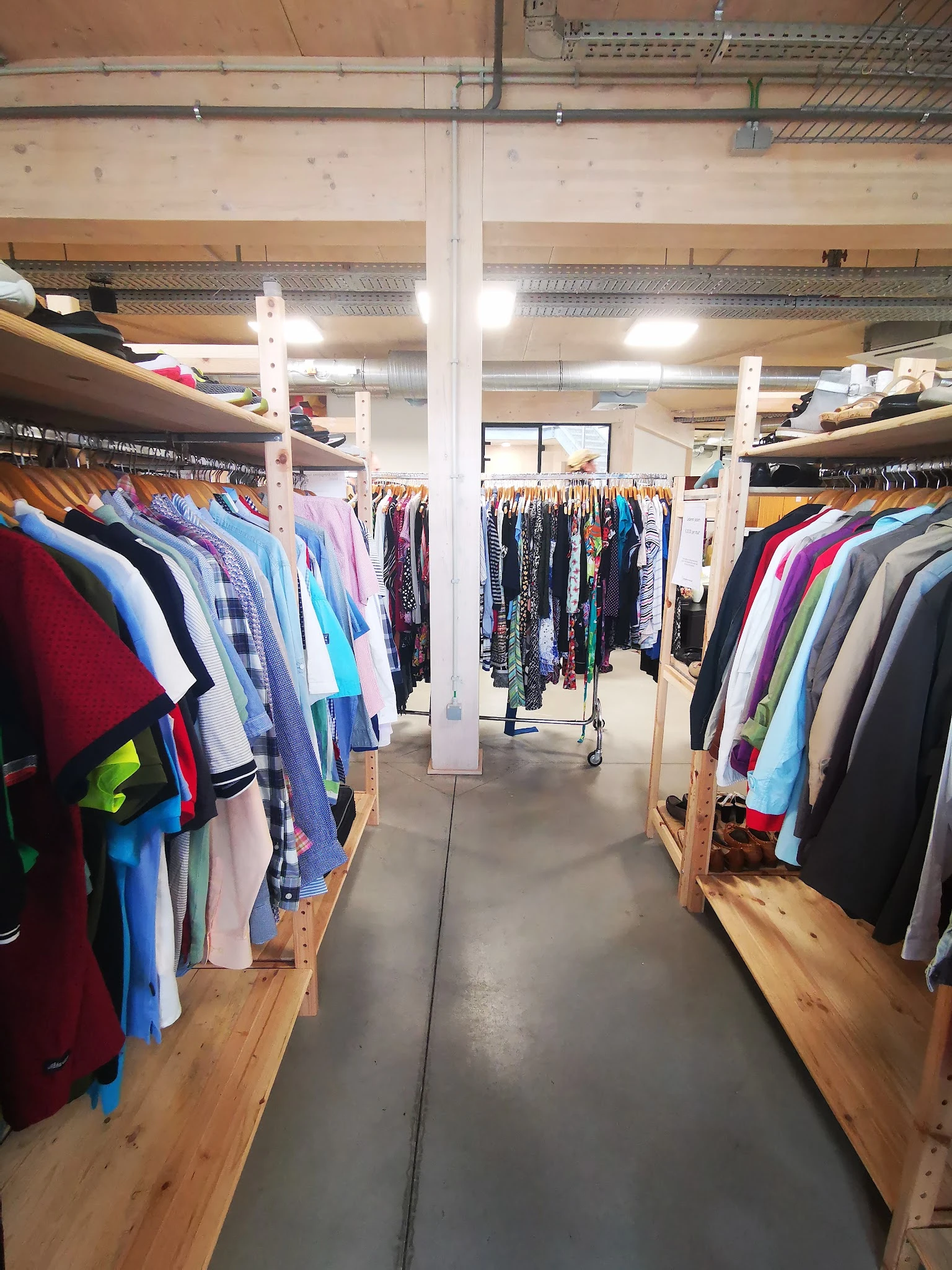 Winkelinterieur met rekken vol tweedehands kleding in verschillende kleuren, witte pilaren en betonnen vloer