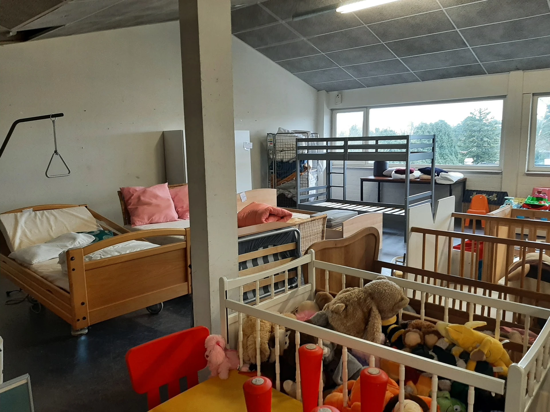 Speelhoek in een kringloopwinkel met babybed, kinderledikantjes, speelgoed en rode stoelen rond een gele tafel