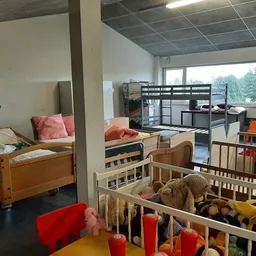 Speelhoek in een kringloopwinkel met babybed, kinderledikantjes, speelgoed en rode stoelen rond een gele tafel