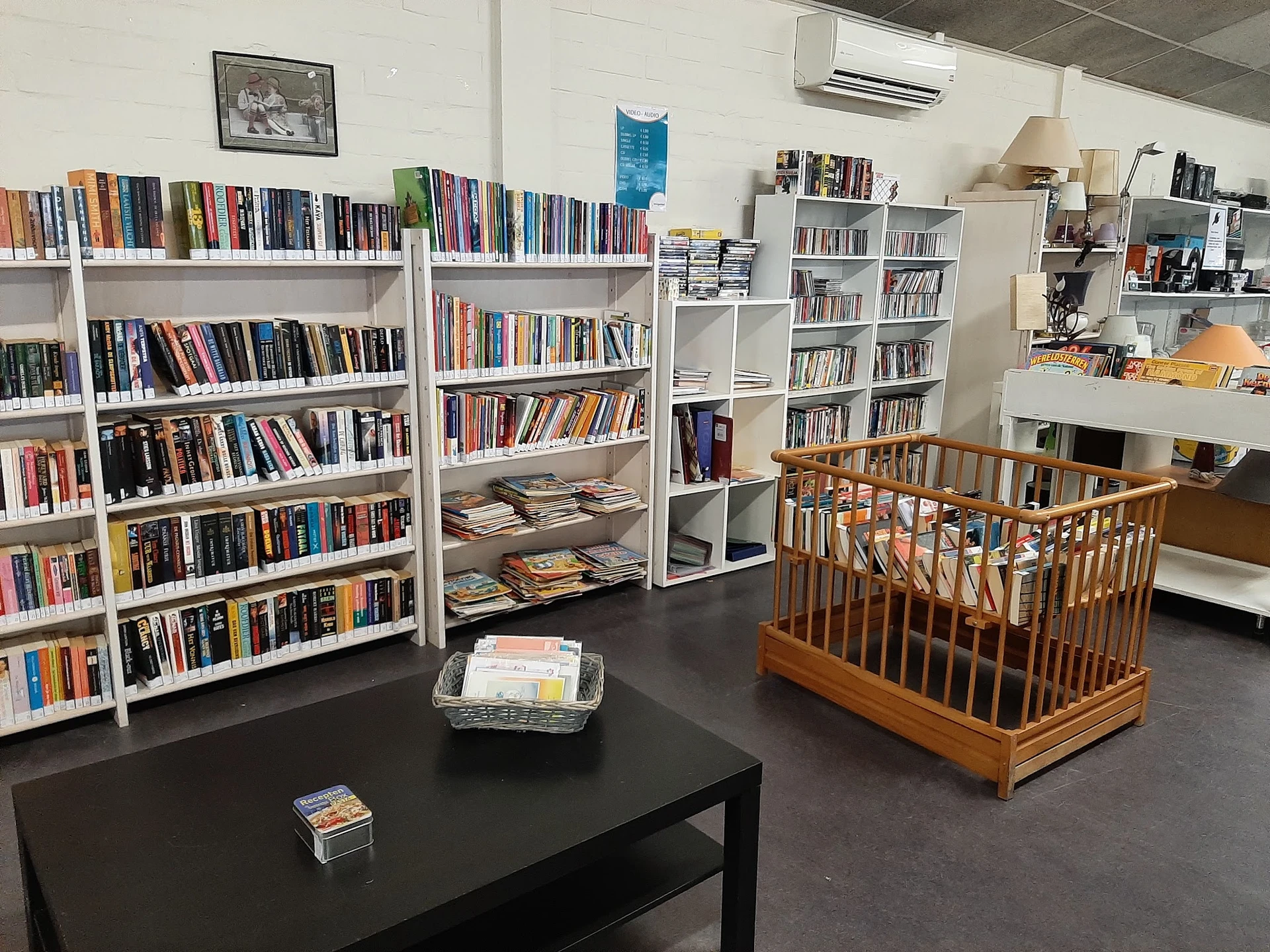 Winkelinterieur met boekenrekken langs de muur, zwarte tafel met items, en houten babybed op grijze vloer