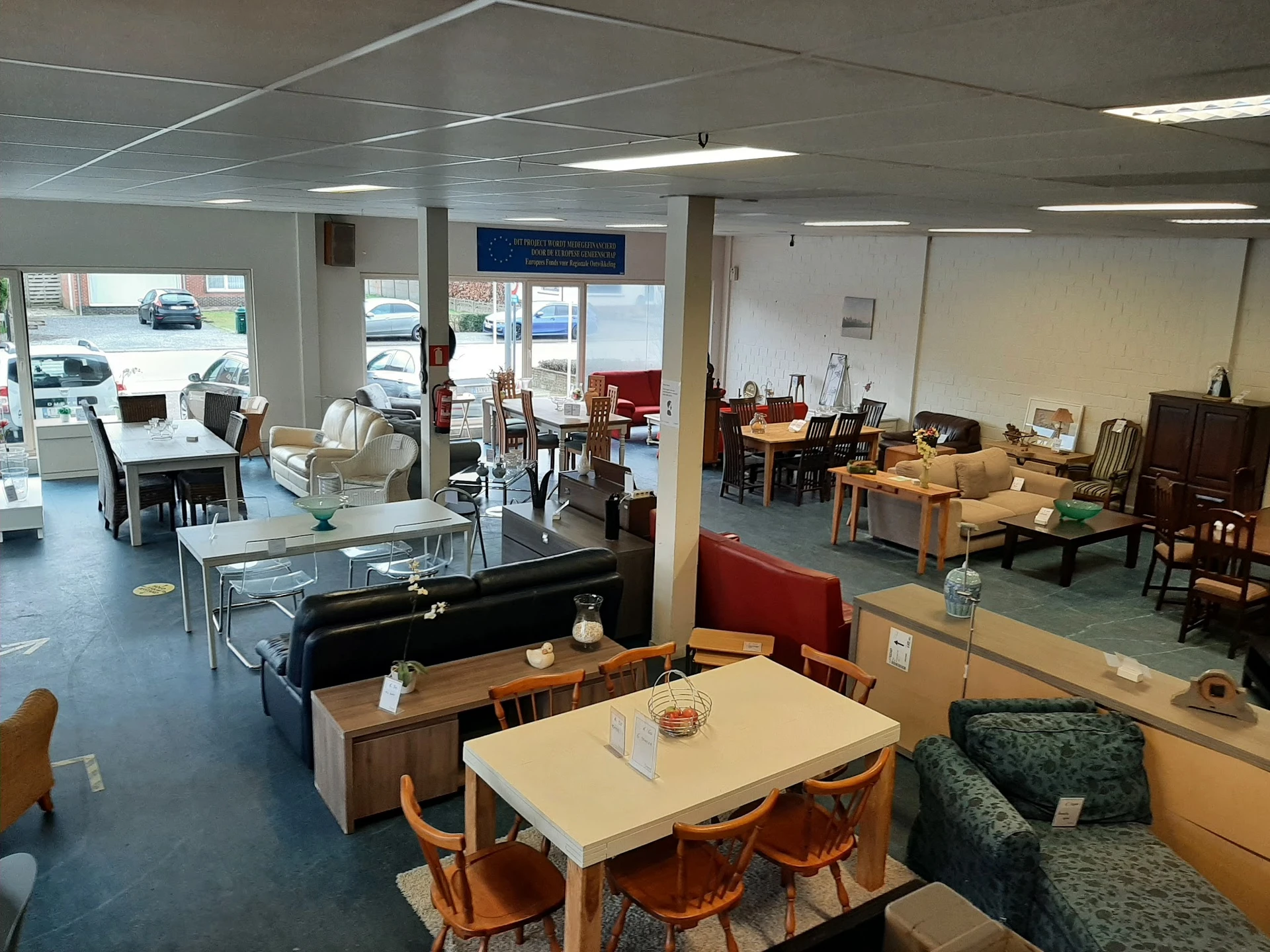 Ruim winkelbinnenruimte met diverse meubels, tafels, stoelen en huisraad uitgestald op blauwe vloer