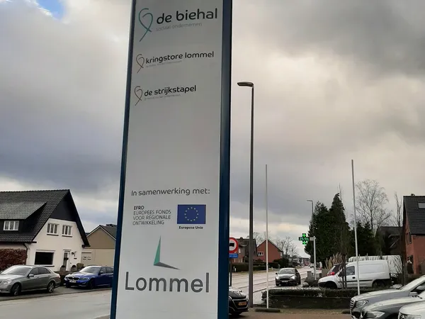 Blauw informatiebord met winkelnaam Lommel en drie aanduidingen voor verschillende afdelingen, staande voor de winkelgevel.
