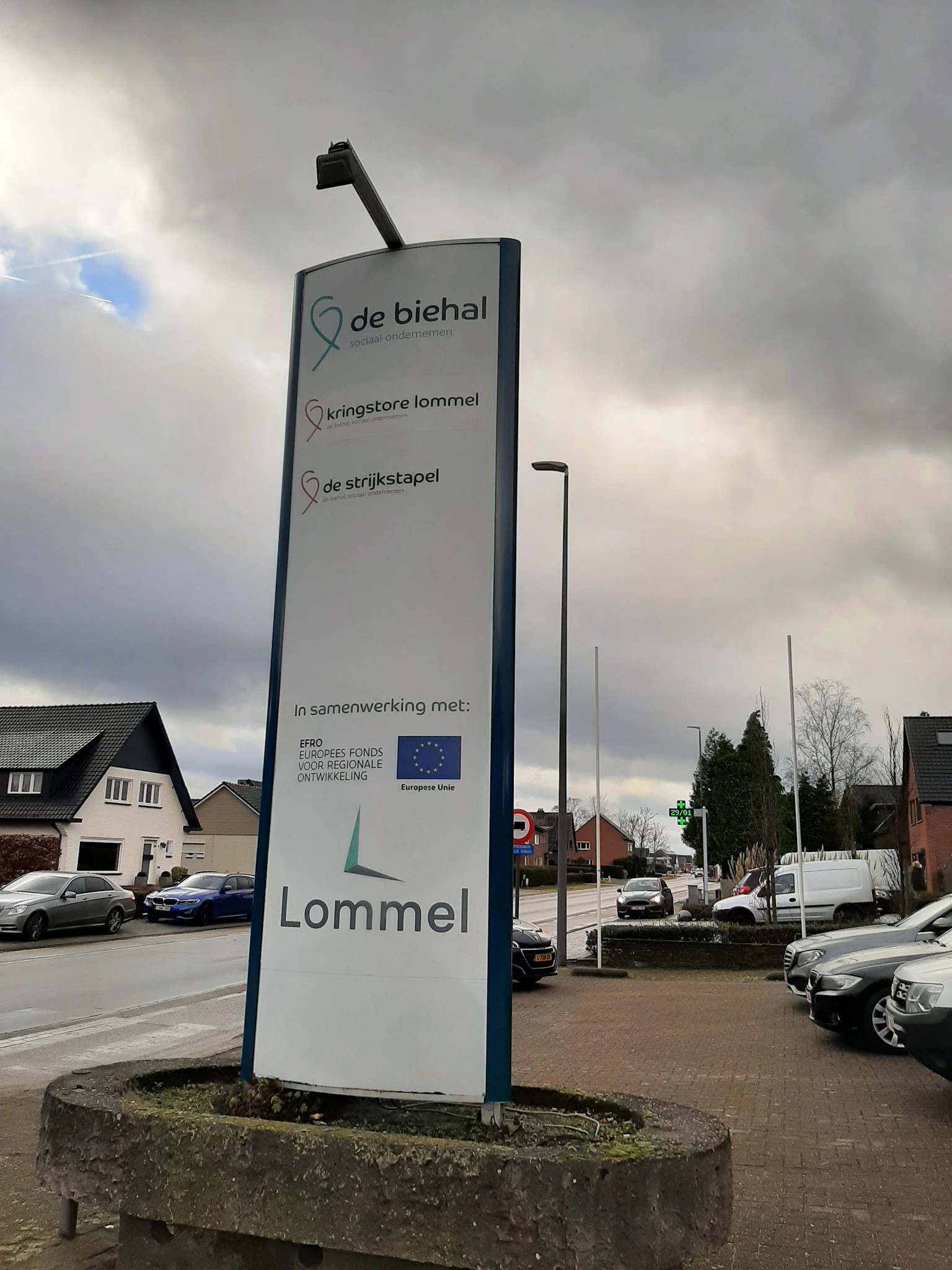 Blauw informatiebord met winkelnaam Lommel en drie aanduidingen voor verschillende afdelingen, staande voor de winkelgevel.