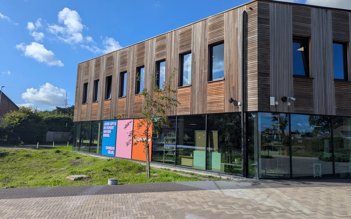 Moderne kringloopwinkel met houten en donkere gevel, grote ramen op begane grond, stellingen met spullen zichtbaar, straatvoo