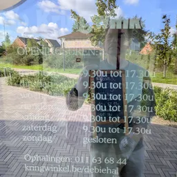 Voorkant van kringloopwinkel met ingang achter paal, terras en groen