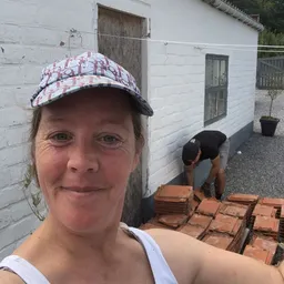 Vrouw met pet poseert voor camera, op achtergrond man die bezig is met tegels