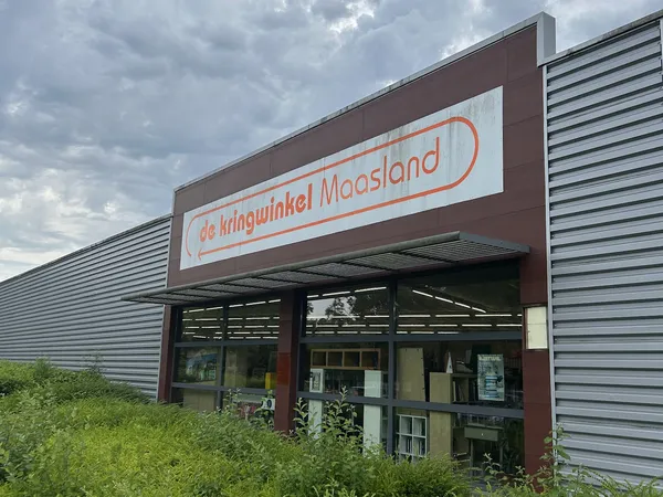 Gevel van Kringwinkel Maasland met bruine pui en wit logo met rode rand, onder bewolkte hemel