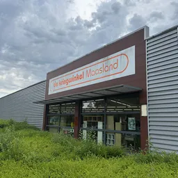 Gevel van Kringwinkel Maasland met bruine pui en wit logo met rode rand, onder bewolkte hemel