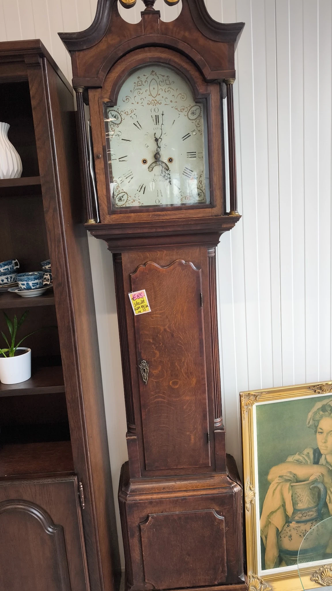 Antieke houten staande klok met glazen kast bovenaan, gele prijssticker zichtbaar