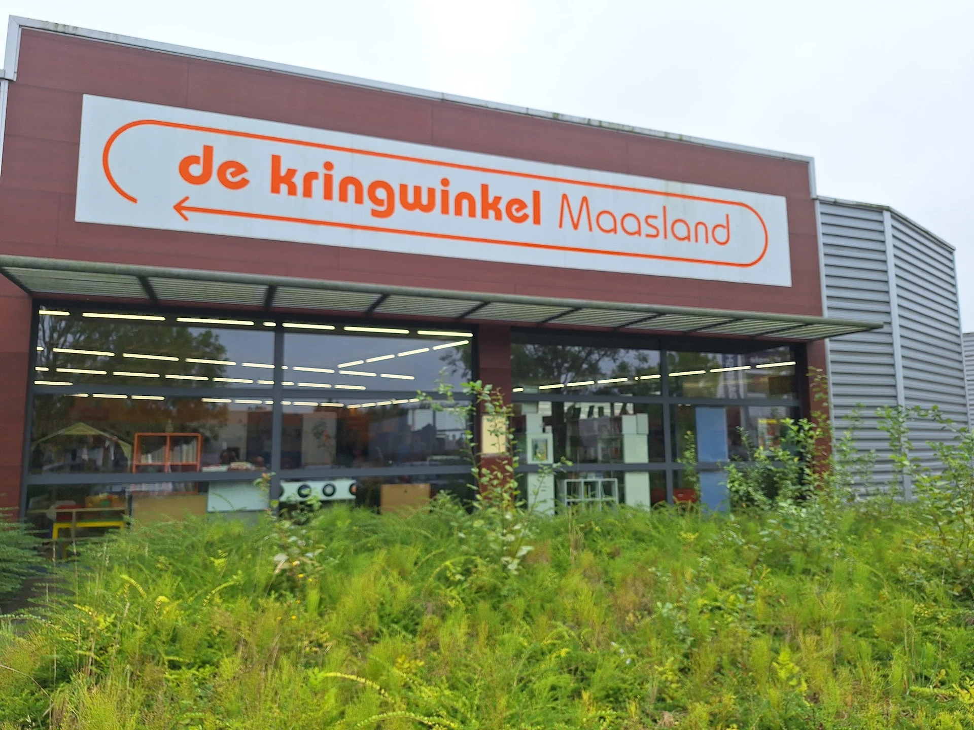Rode gevel van kringloopwinkel met wit logo 'de kringwinkel Maasland', grote ramen en groeiend onkruid ervoor
