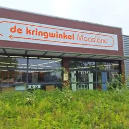 Rode gevel van kringloopwinkel met wit logo 'de kringwinkel Maasland', grote ramen en groeiend onkruid ervoor