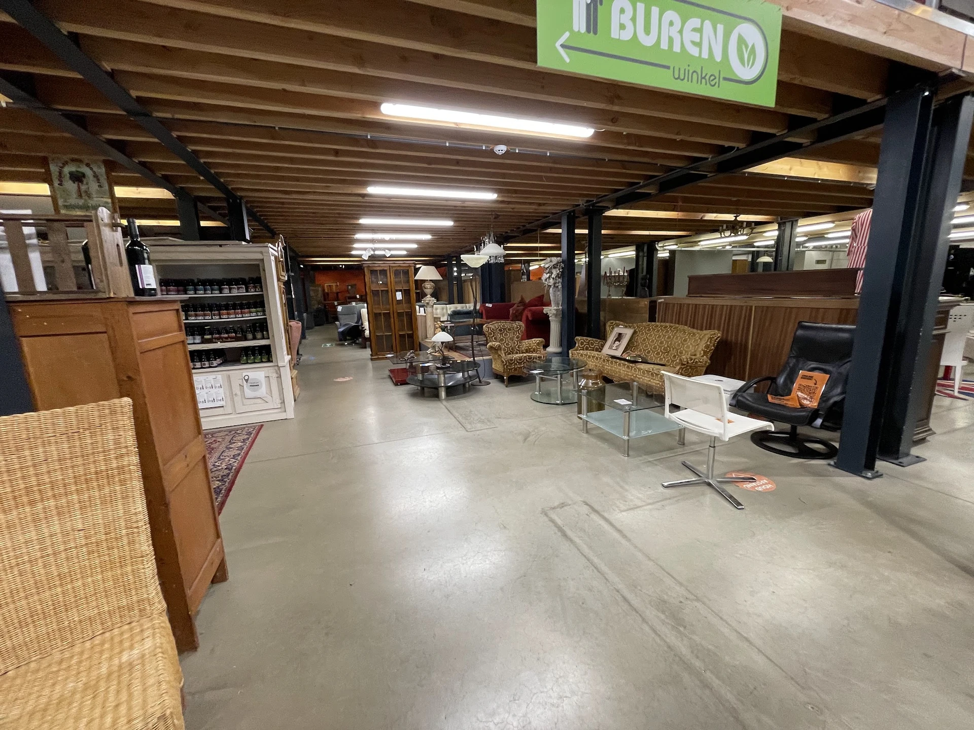 Winkelinterieur met diverse meubelen, rekken en spullen op betonvloer onder houten balkenplafond