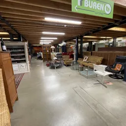 Winkelinterieur met diverse meubelen, rekken en spullen op betonvloer onder houten balkenplafond