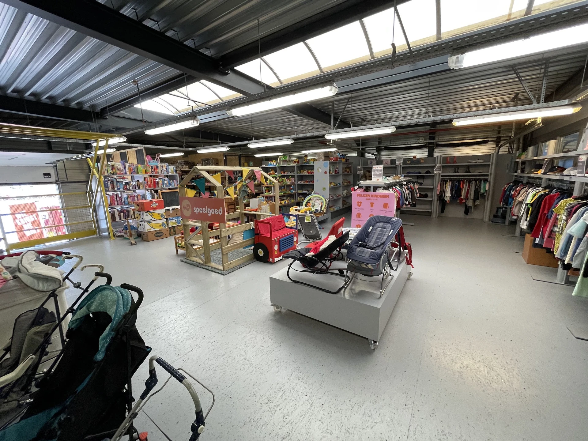 Ruime winkelhal met stellingen, kleding aan rekken, speelgoed en diverse spullen op tafels