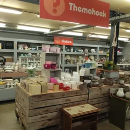 Winkelvloer met houten display-tafels vol huishoudwaren, stellingen op de achtergrond, rode Themahoek-bord zichtbaar