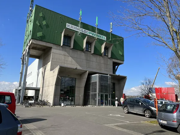 Moderne gevel van kringloopwinkel met groene bovenbouw en betonnen onderdeel, parkeerplaats met auto's ervoor