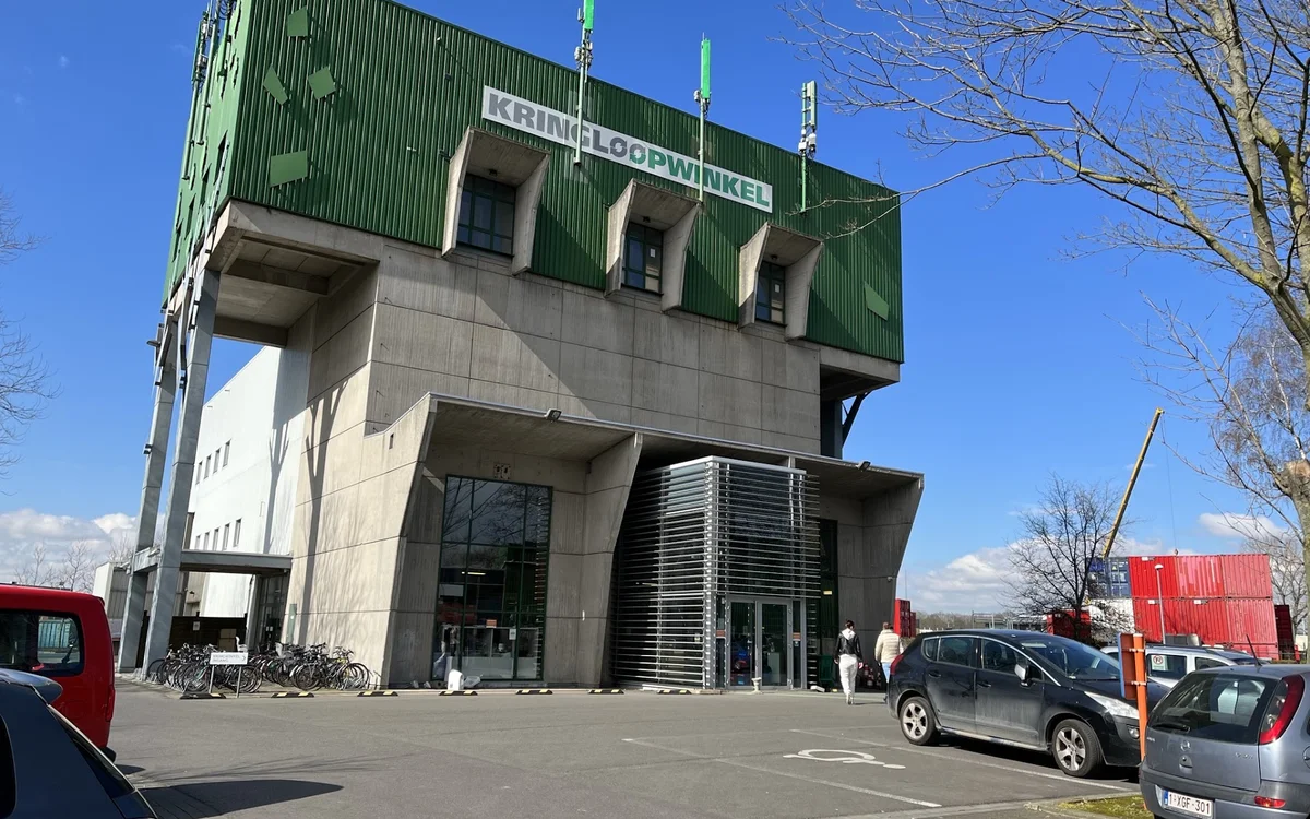 Moderne gevel van kringloopwinkel met groene bovenbouw en betonnen onderdeel, parkeerplaats met auto's ervoor