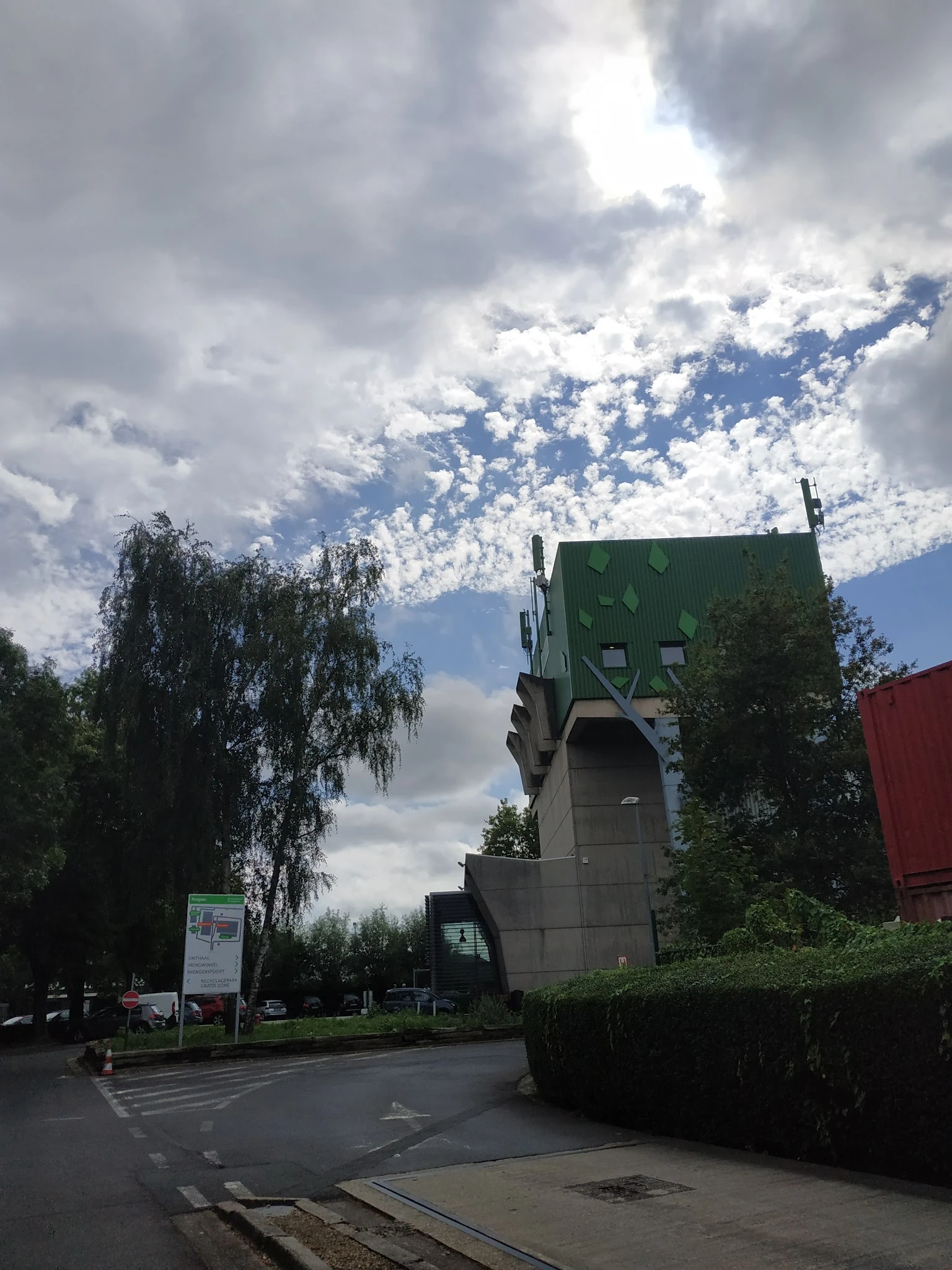 Groen dak met ramen en grijze muur van een gebouw, omgeven door bomen en bewolkte hemel