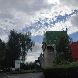 Groen dak met ramen en grijze muur van een gebouw, omgeven door bomen en bewolkte hemel