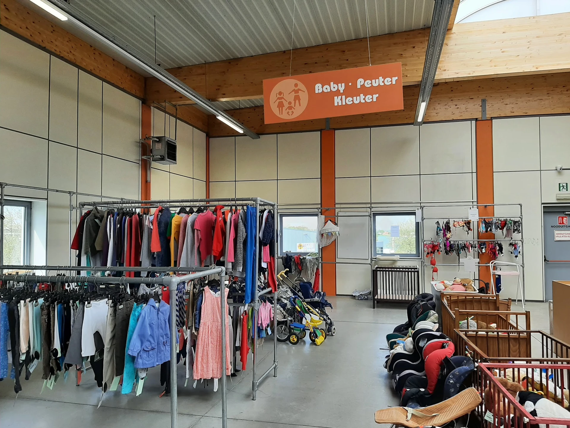 Winkelinterieur met rekken babykleding, kinderwagen en speelgoed onder oranje-wit signage