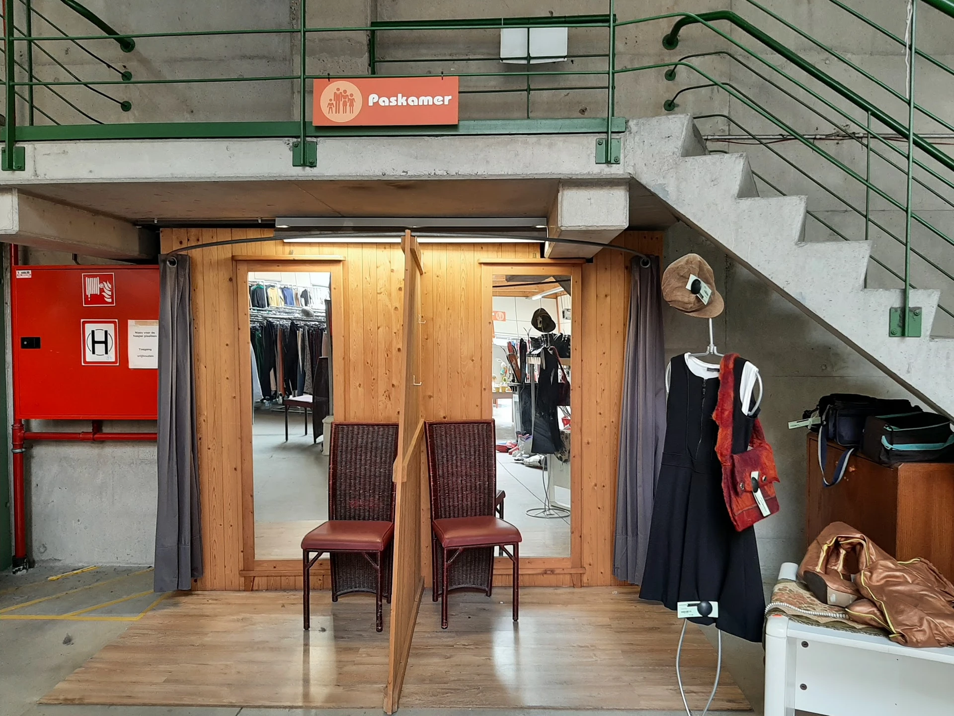 Winkelinterieur met houten deur, bruine stoelen, kleding op hangers en stellingen onder trap