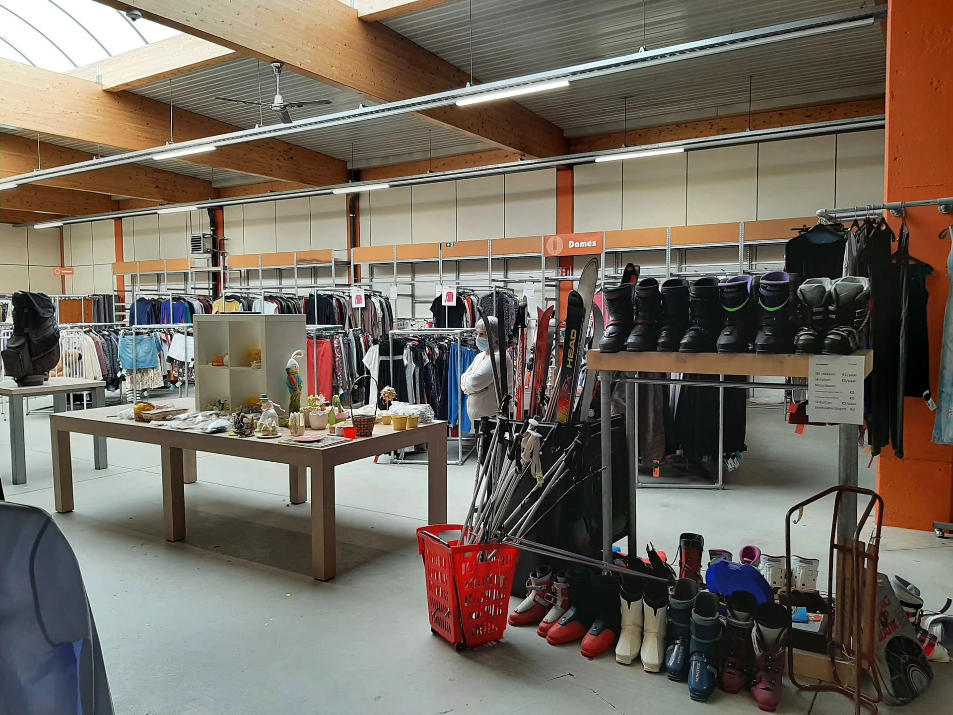 Winkelinterieur met kledingrekken, stellingen met zwarte tassen, bruine tafel in het midden en rode mand met schoenen op de v