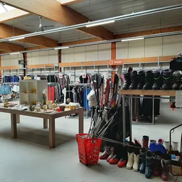 Winkelinterieur met kledingrekken, stellingen met zwarte tassen, bruine tafel in het midden en rode mand met schoenen op de v
