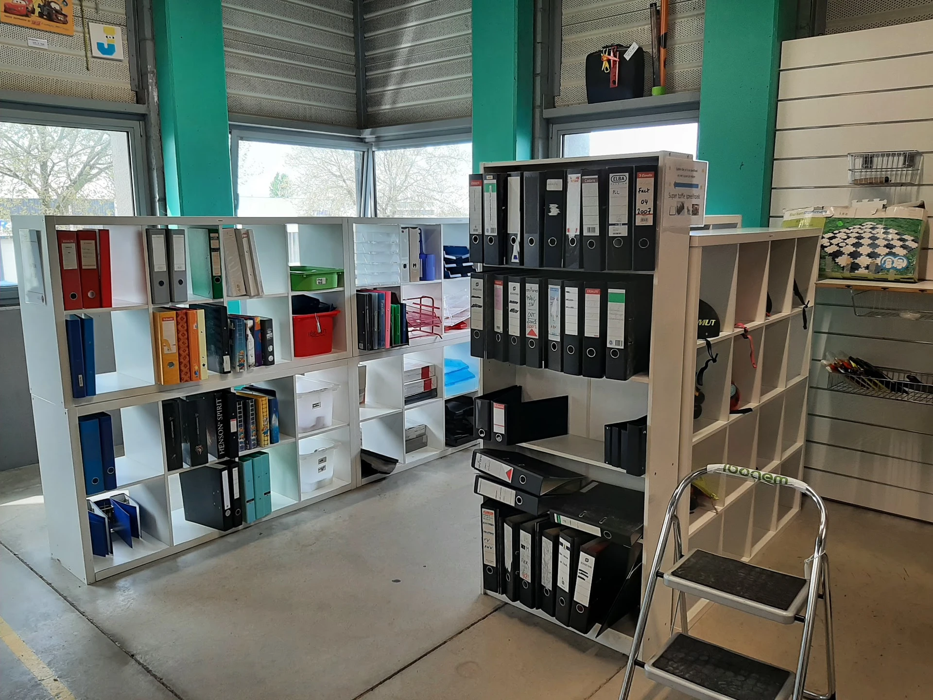 Winkelvloer met witte stellingen vol boeken, ordners en kantoorartikelen, groene zuilen en ramen op de achtergrond