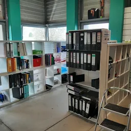 Winkelvloer met witte stellingen vol boeken, ordners en kantoorartikelen, groene zuilen en ramen op de achtergrond
