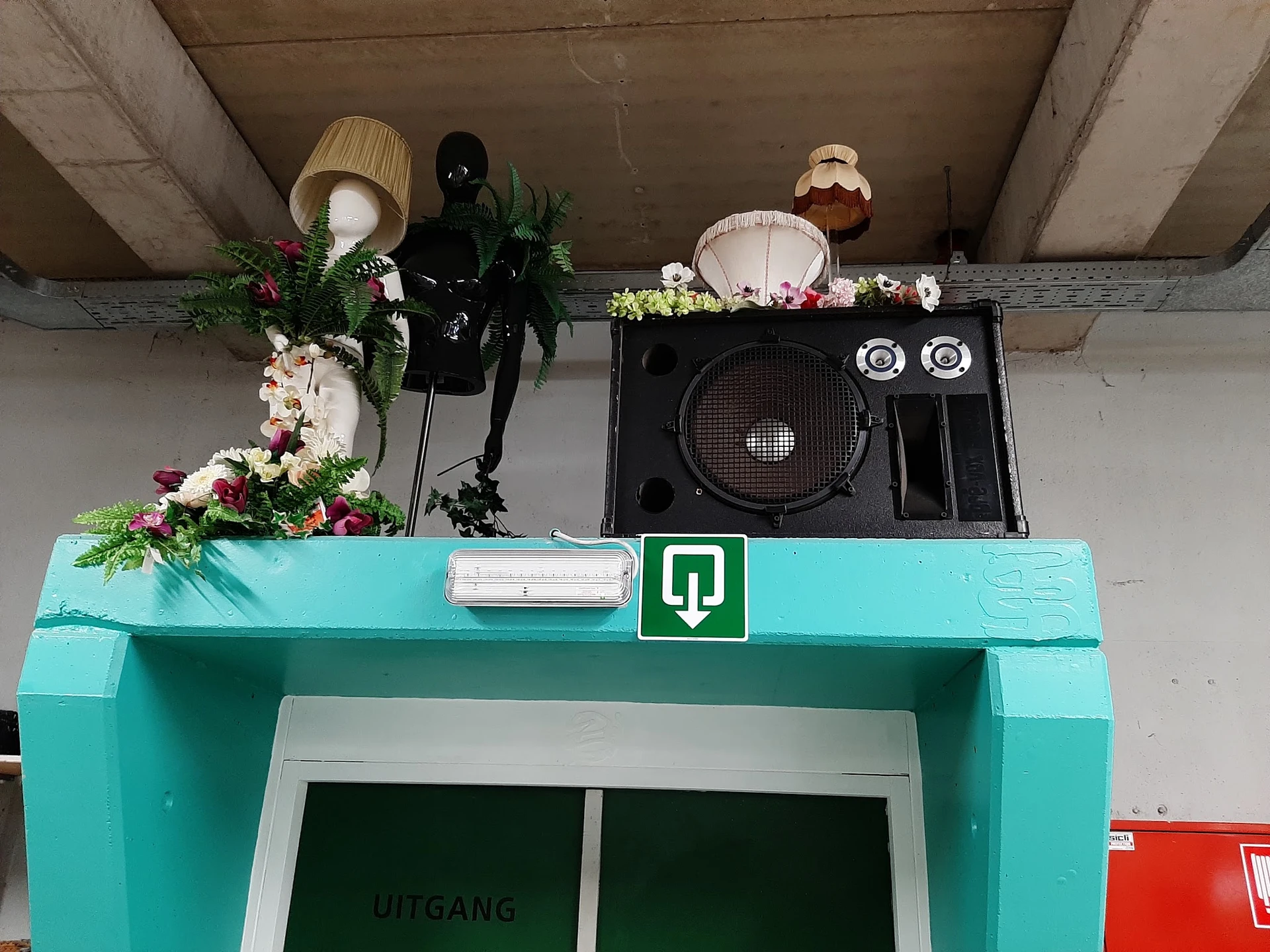 Turquoise winkelingang met groene deur en decoratieve items op het dak, waaronder speakers en kunstbloemen