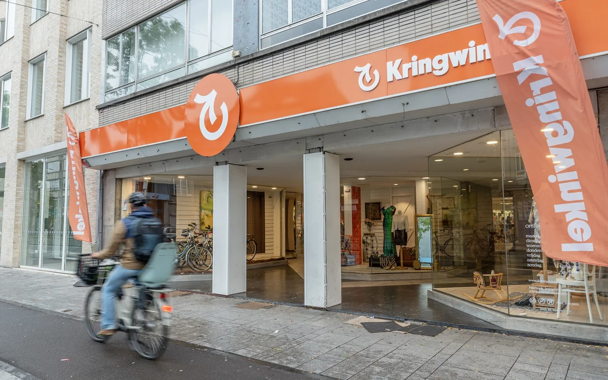 Oranje gevel van kringloopwinkel met logo en grote ramen, fietser in beweging op straat
