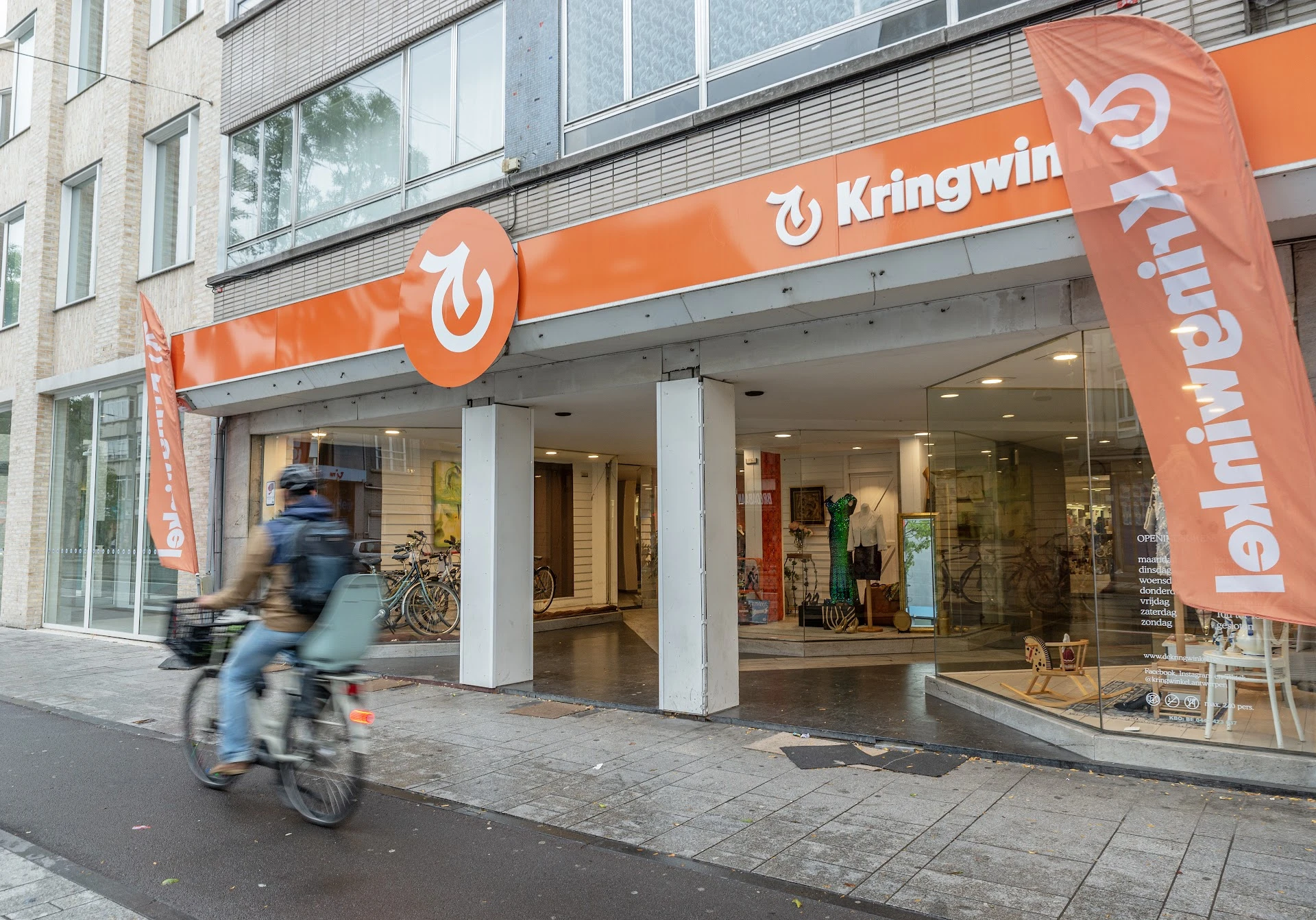 Oranje gevel van kringloopwinkel met logo en grote ramen, fietser in beweging op straat