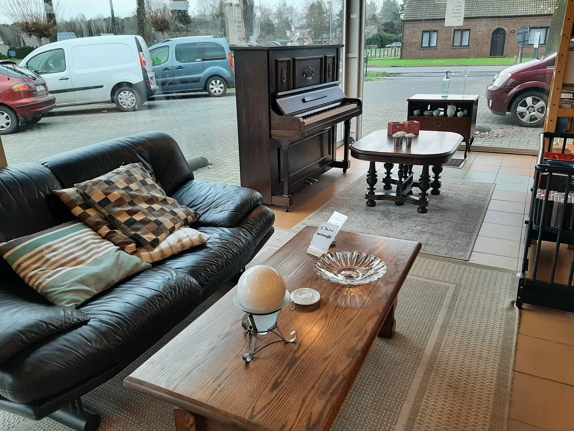 Winkelvloer met zwarte leren bank, houten salontafel, piano en verschillende meubelstukken; ramen tonen parkeerplaats buiten