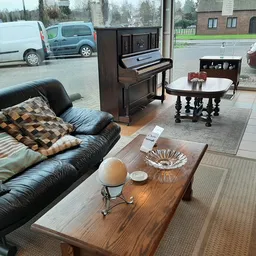 Winkelvloer met zwarte leren bank, houten salontafel, piano en verschillende meubelstukken; ramen tonen parkeerplaats buiten