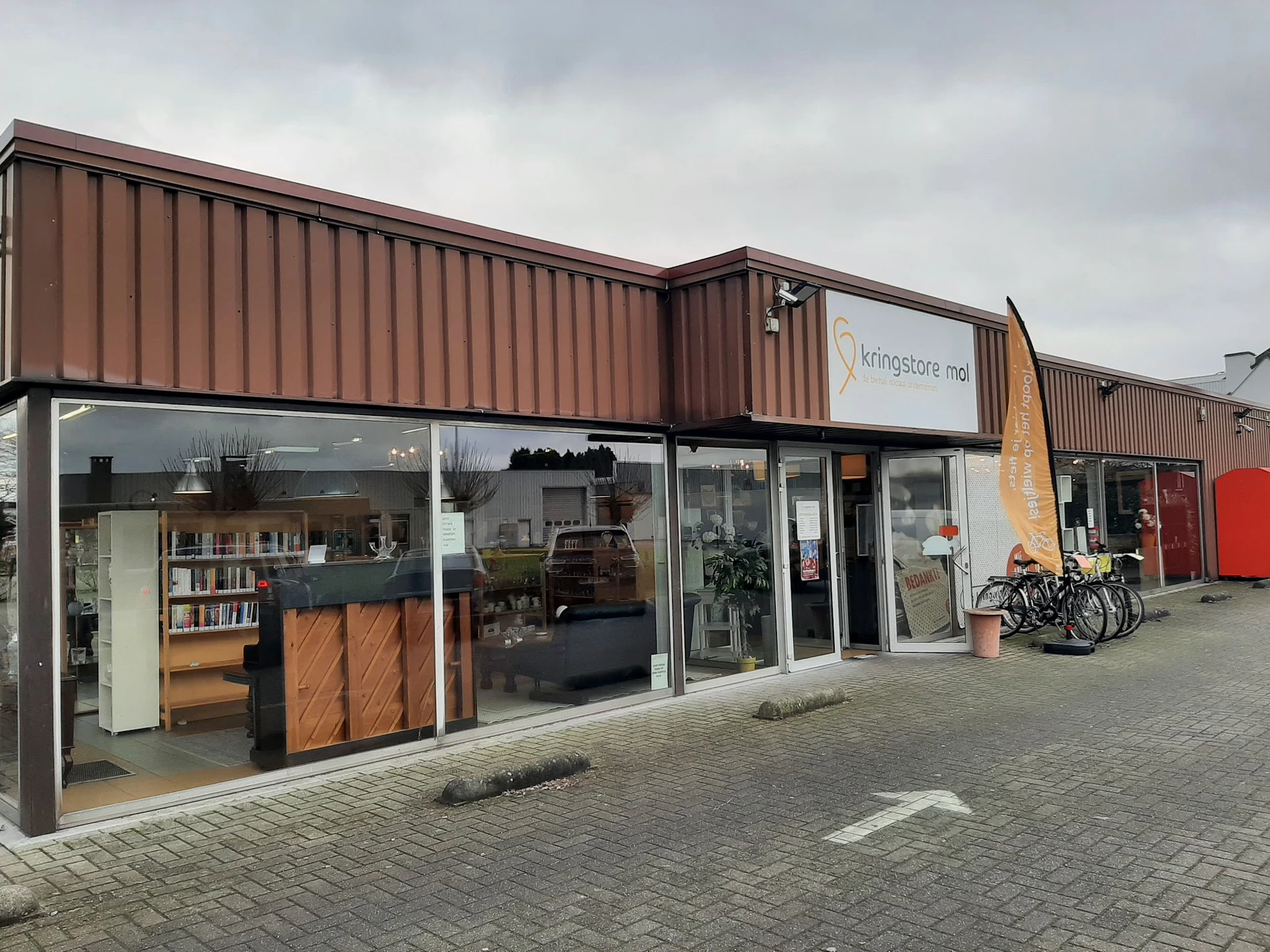 Voorgevel van kringloopwinkel met bruine golfplaten, glasdeuren, etalage met meubels en fietsen ernaast