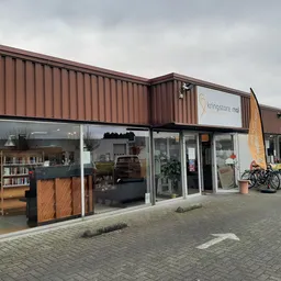 Voorgevel van kringloopwinkel met bruine golfplaten, glasdeuren, etalage met meubels en fietsen ernaast