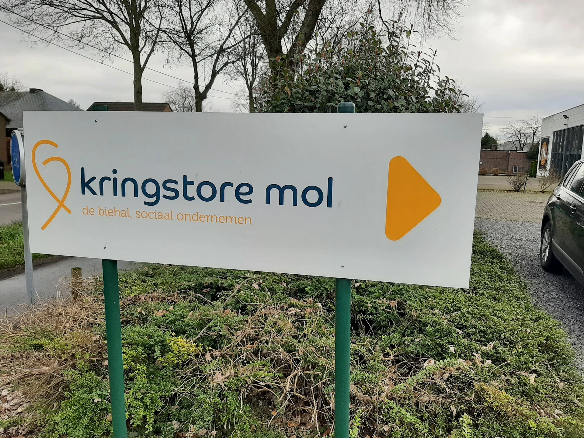 Wit bord met logo en naam van kringloopwinkel aan ingang, groen onderhoud eronder