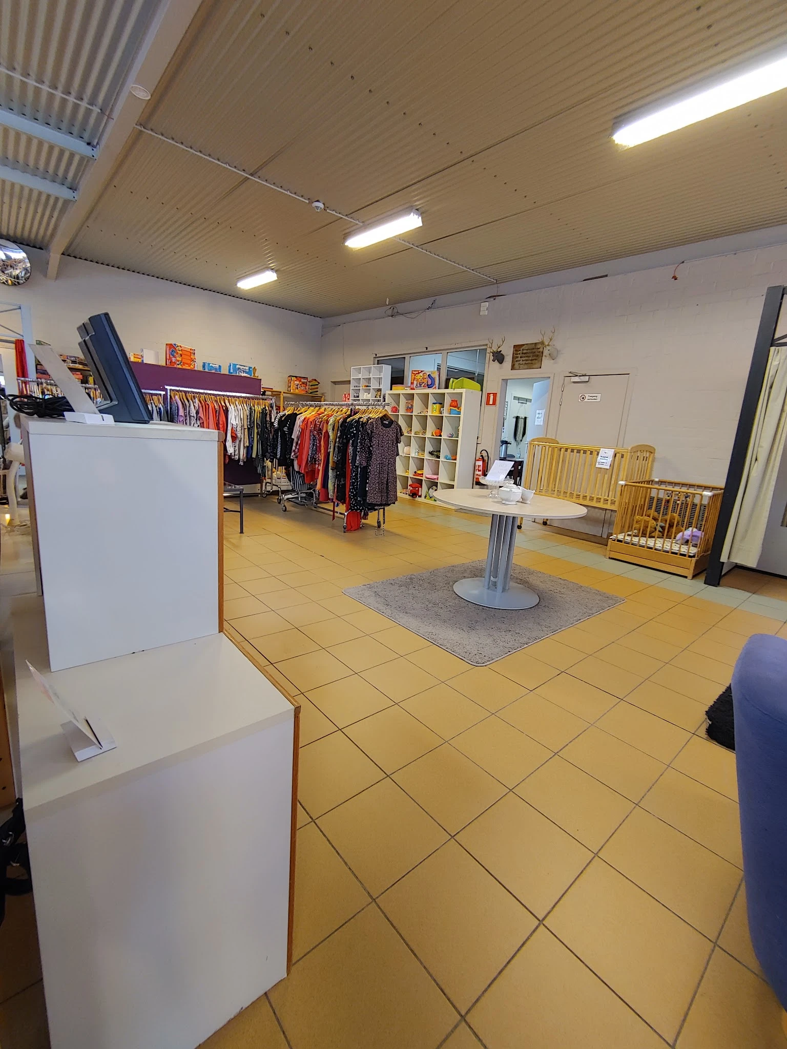 Winkelvloer van kringloopwinkel met kleding op rekken, stellingen, gele tegelvloer en grijze loopmat in het midden