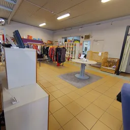 Winkelvloer van kringloopwinkel met kleding op rekken, stellingen, gele tegelvloer en grijze loopmat in het midden