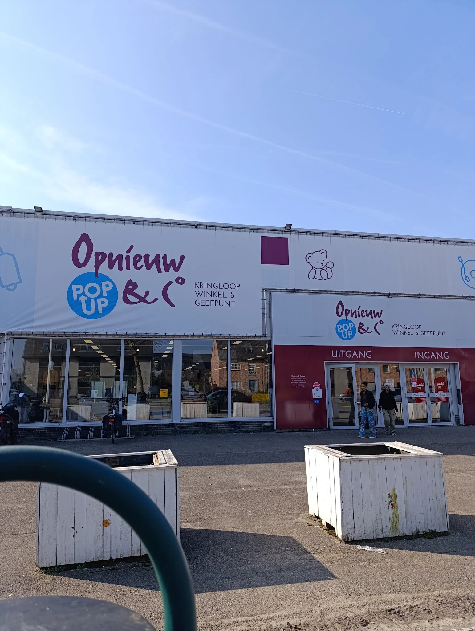 Gevel van kringloopwinkel met wit en bordeauxrood geschilderde pui, grote ramen, winkelnaam zichtbaar, ingang en display aan 