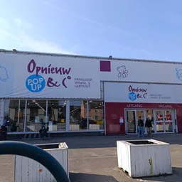 Gevel van kringloopwinkel met wit en bordeauxrood geschilderde pui, grote ramen, winkelnaam zichtbaar, ingang en display aan 