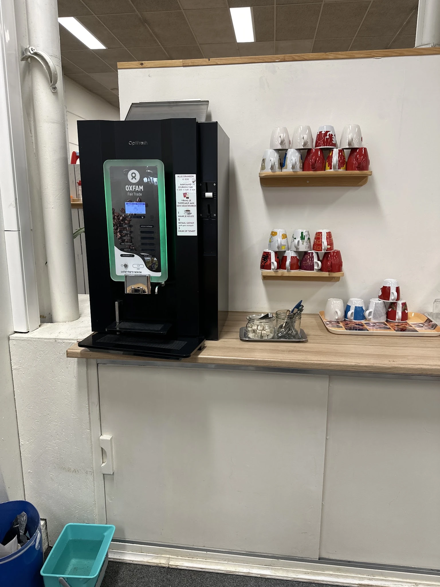 Hoek van kringloopwinkel met koffieapparaat, houten schappen met potjes en cosmeticaproducten, en houten werktafel