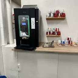 Hoek van kringloopwinkel met koffieapparaat, houten schappen met potjes en cosmeticaproducten, en houten werktafel