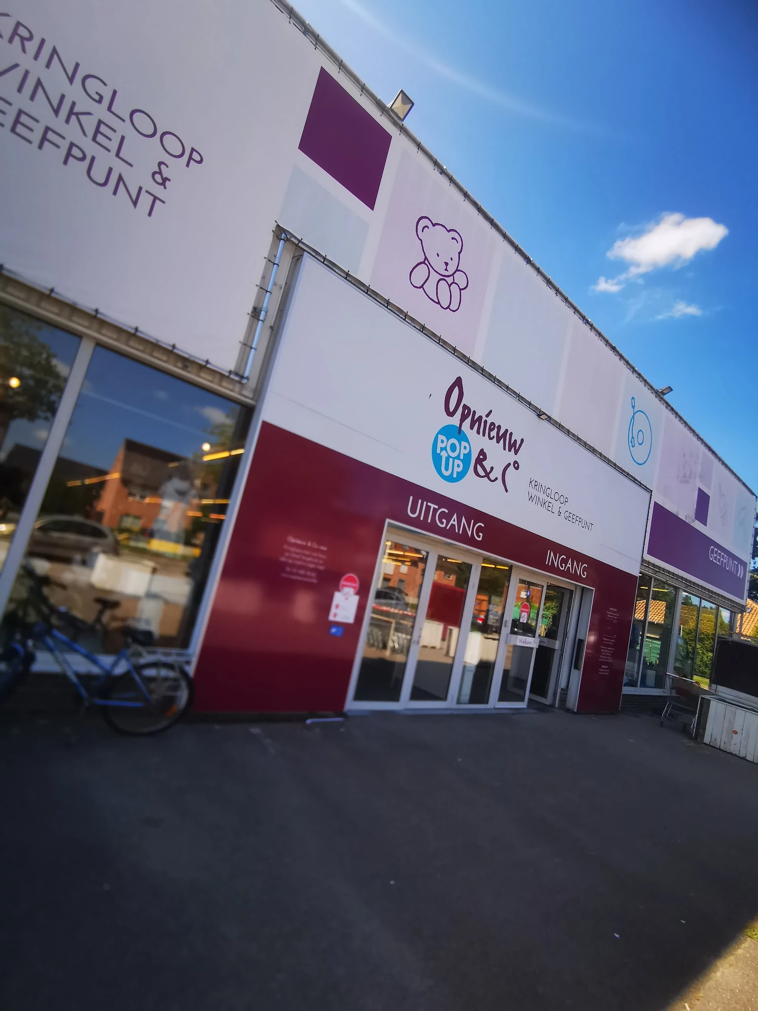 Gevel van kringloopwinkel met witte en bordeauxrode bekeding, winkelnaam Opnieuw & Co zichtbaar, grote etalageruiten en ingan