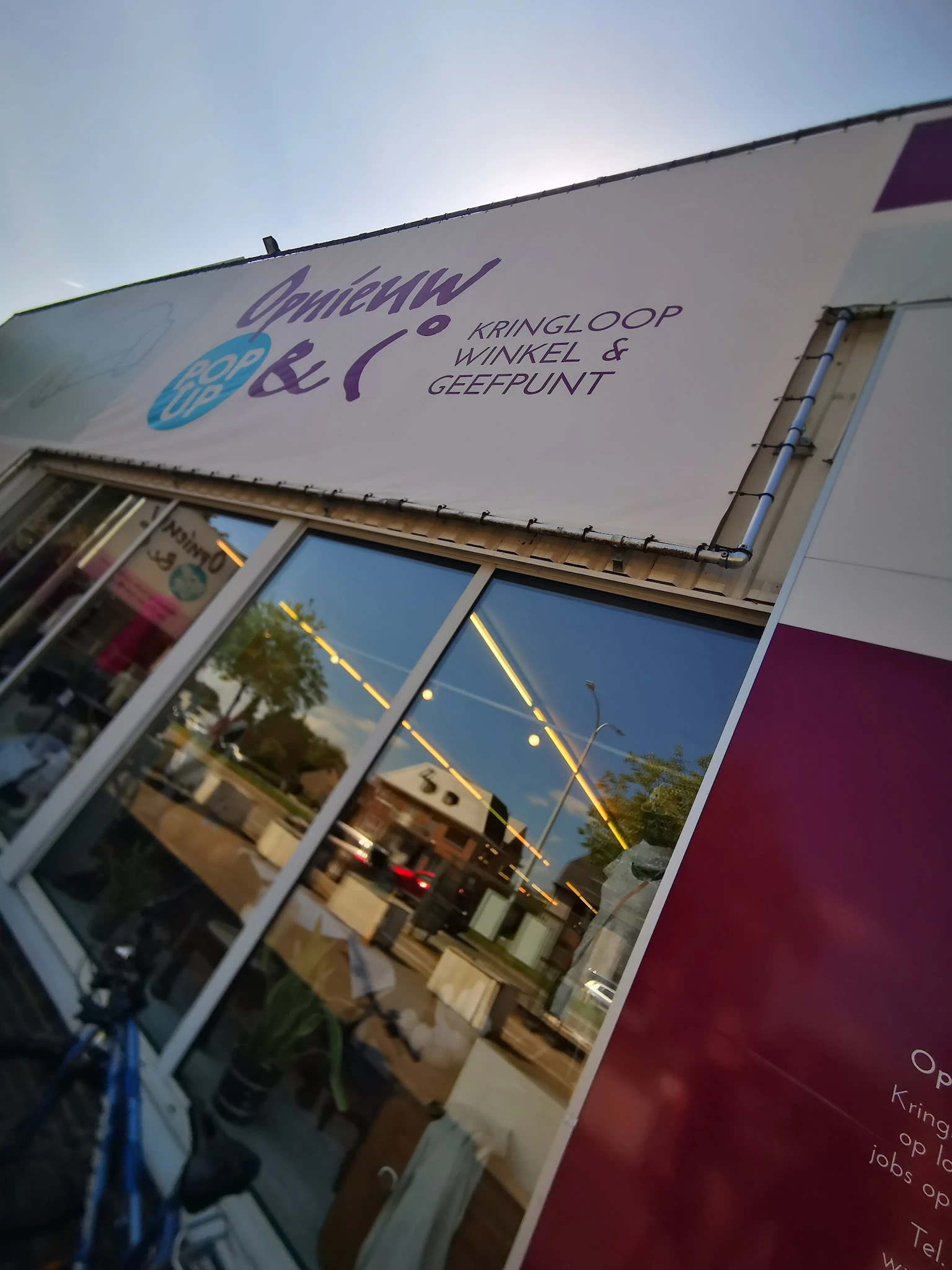 Gevel van kringloopwinkel met winkelnaam en logo op zijlicht, straatreflex in raam zichtbaar