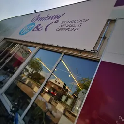 Gevel van kringloopwinkel met winkelnaam en logo op zijlicht, straatreflex in raam zichtbaar