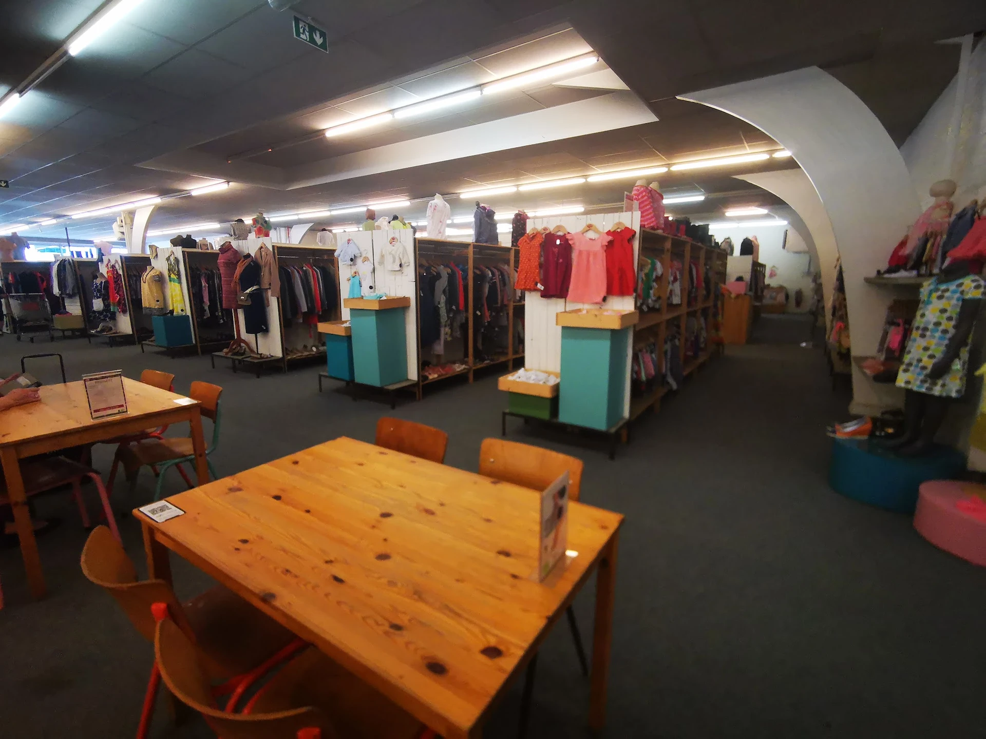 Winkelvloer met houten tafels, oranje stoelen en kledingrekken met diverse artikelen in een kringloopwinkel