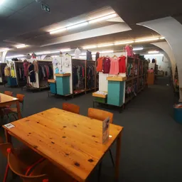 Winkelvloer met houten tafels, oranje stoelen en kledingrekken met diverse artikelen in een kringloopwinkel