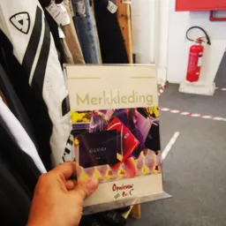 Hand met een merkkleding-catalogus of brochure met Gucci-afbeelding, in een kringloopwinkel