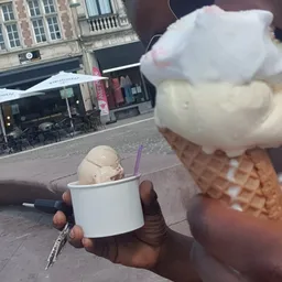 Hand met twee ijsjes (een in een beker, een in een wafel) op een terras met winkelstraat op de achtergrond